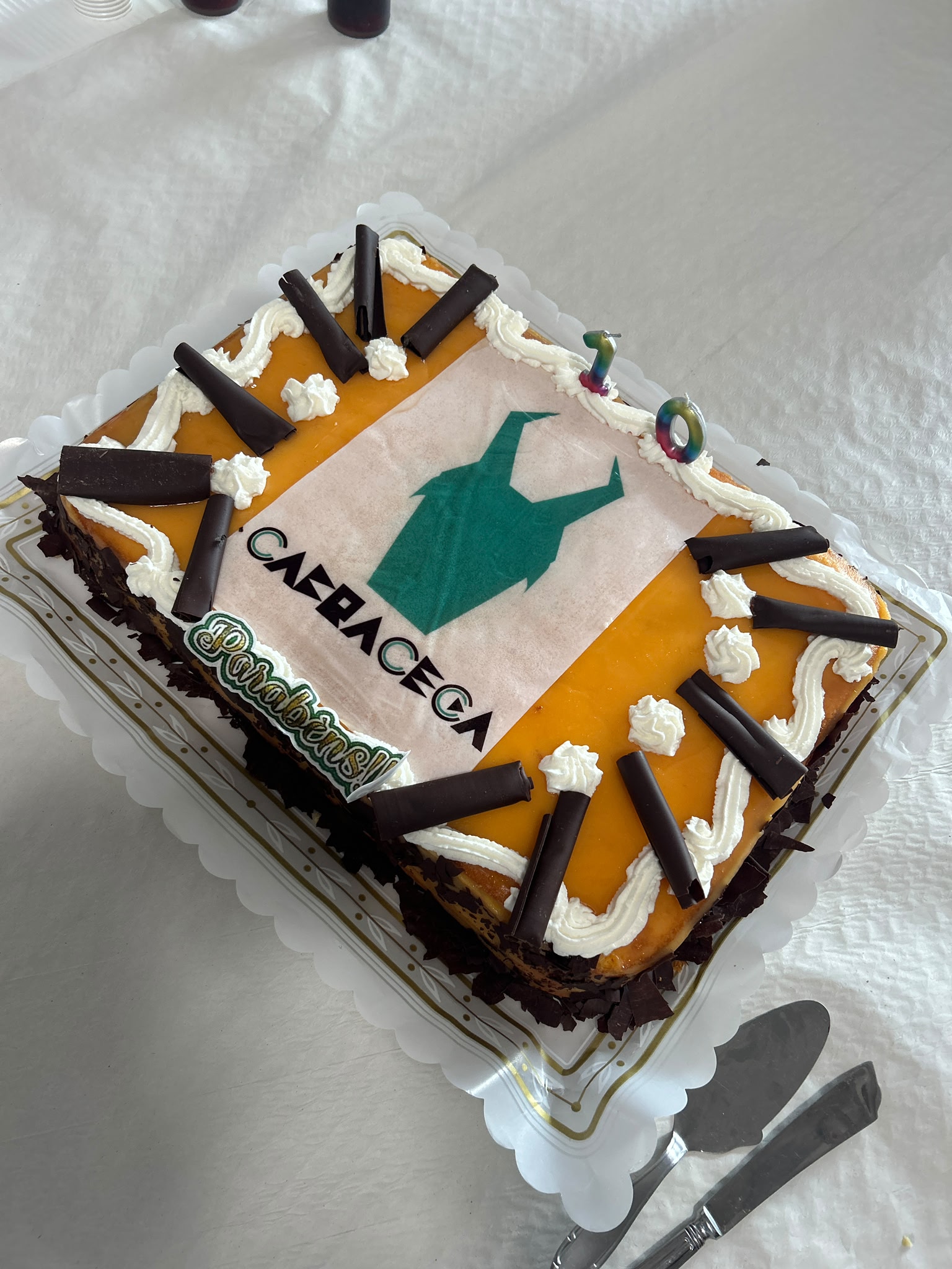 Um bolo retangular decorado com cobertura amarela e chantilly branco. No centro há uma folha com desenho de uma cabeça de uma cabra verde com dois “chifres” e, por baixo, o texto em preto “CABRA CEGA” (a parte do meio da palavra “CAÇA” está parcialmente coberta pelo brilho). Perto de um dos cantos está a palavra “Parabéns!!” em verde contornada de branco. Em volta da borda superior do bolo há uma faixa de chantilly branco em zigue?zague, com vários rolinhos de chocolate escuro apoiados sobre o chantilly. Na parte superior direita do bolo há duas velas em forma de número, “1” e “0”, coloridas em degradê, indicando 10 anos. Os lados do bolo têm raspas grandes de chocolate escuro. O bolo está sobre uma base de plástico branco com detalhes dourados, colocada numa toalha clara.